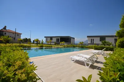 Luxury Outdoor Swimming Pool and Sun Deck. A bright, modern outdoor swimming pool area with a sparkling blue pool, surrounded by light-colored decking and green landscaping. White lounge chairs line the pool, offering a relaxing amenity.