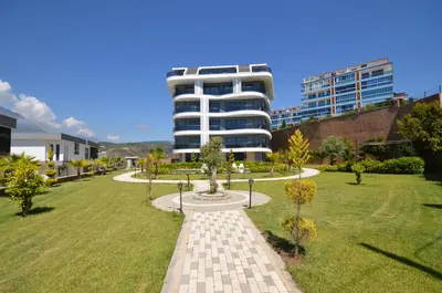 Modern Residential Building with Lush Green Garden. Exterior view of a modern residential building featuring curved balconies, set within a lush, well-maintained green garden with a paved pathway, under a clear blue sky.