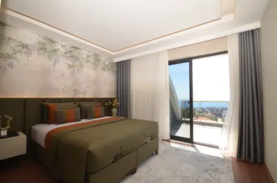 Stylish Bedroom with Balcony City and Sea Views. Modern bedroom features a green bed, botanical wallpaper, and wood floor. Large glass door opens to a balcony revealing city and sea views, providing bright natural light.