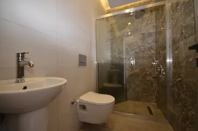Modern Bathroom with Elegant Walk-in Shower and Sink. This modern bathroom features a white pedestal sink, a wall-mounted toilet, and a spacious glass-enclosed walk-in shower with stylish brown marble-look tiles on the walls.