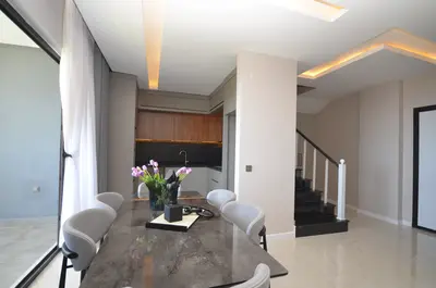 Modern Open-Plan Living Room Dining Kitchen Area. Modern open-plan living and dining area with a stylish dark marble table and grey chairs. Features include an integrated kitchen, ambient ceiling lighting, and a large sliding glass door. A modern staircase is visible.