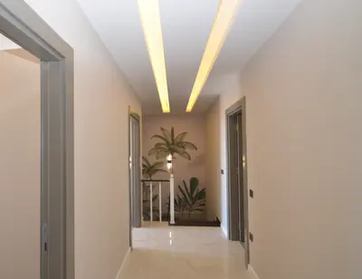 Modern Hallway with Recessed Lighting and Decorative Mural. A modern, well-lit hallway with glossy tiled flooring, light walls, and elegant recessed ceiling lights. A decorative palm tree mural at the end adds character above a staircase with a white balustrade.