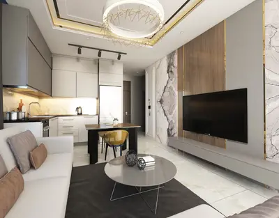 Stylish Open Plan Living Area and Kitchen. Modern open-concept living room and kitchen with sleek grey cabinets, white sofa, and a wall-mounted TV on a marble-paneled wall. Features stylish contemporary lighting and light tiled flooring.