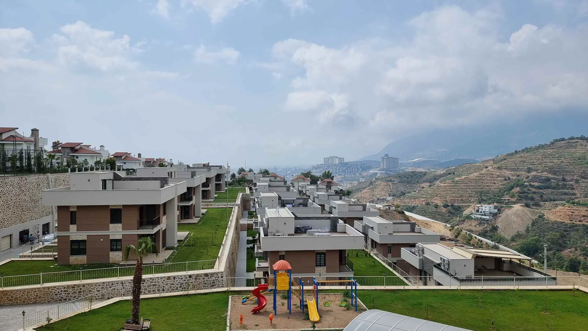 Vile na padini s dječjim igralištem i pogledom na grad. Moderni stambeni kompleks s novim vilama na padini i zelenim travnjacima. Sadrži šareno dječje igralište, s pogledom na udaljeni grad i planine pod oblačnim nebom.