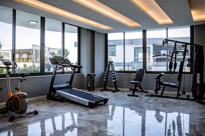 Modern Private Gym with Panoramic Windows and Full Equipment. A modern, well-equipped private gym with a treadmill, exercise bike, weights, and multi-station machine. Features sleek marble-patterned flooring, bright ceiling lights, and large panoramic windows offering exterior views.