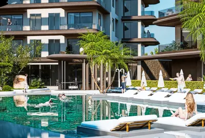 Luxury Outdoor Swimming Pool and Modern Residence. An expansive, modern outdoor swimming pool at a luxury residential complex. Features clear turquoise water, stylish white sun loungers, lush palm trees, and people enjoying the amenities.