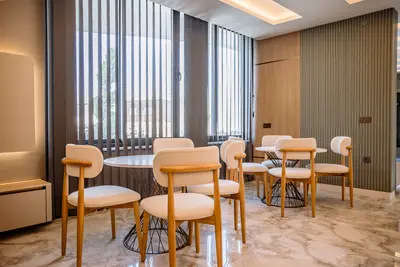 Elegant Dining Area with Marble Floors and Natural Light. A modern dining area with glossy marble floors, featuring two round tables and stylish cream chairs. Large windows with vertical blinds provide natural light, complemented by elegant ceiling and wall lighting.