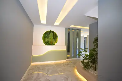Modern Lobby with Green Wall and Integrated Lighting. A modern entrance or lobby with elegant marble flooring and bright recessed ceiling lights. It features a striking circular green wall, a stylish wave-design desk, and an integrated planter with greenery.