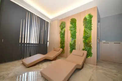 Luxurious Relaxation Room with Vertical Garden Feature. A modern spa-like relaxation room featuring two comfortable beige lounge chairs, a striking vertical plant wall, and polished marble flooring under soft cove lighting. Includes a window with vertical blinds.
