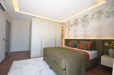 Modern Bedroom with Built-in Wardrobe and Botanical Mural. A modern bedroom with warm hardwood floors and a white rug. Features a large bed with a green headboard and botanical mural, built-in white wardrobe, and cove ceiling lighting. Includes air conditioning.