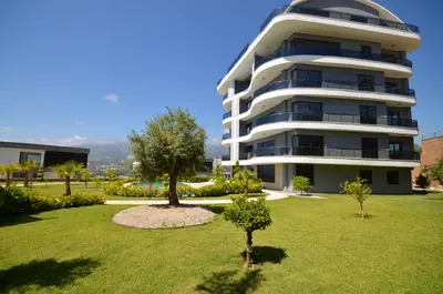 Luxury Modern Apartment Building with Landscaped Garden and Pool. Modern residential building exterior with curved balconies, grey facade. Features a lush green garden, inviting swimming pool, and scenic distant mountains under a clear sky.