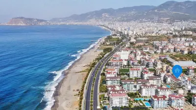 Panoramic Alanya Coastal City View with Beach. Aerial view of a vibrant Turkish coastal city, showcasing a long sandy beach, clear blue sea, and bustling road. Modern residential buildings with pools line the scenic urban landscape against mountains.