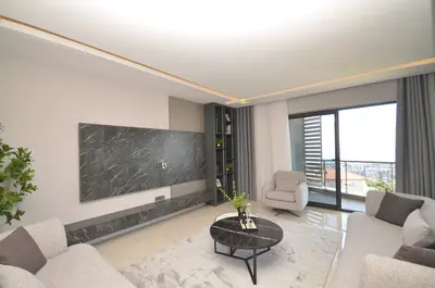 Modern Living Room with Stylish Furnishings and City View. A modern, elegantly furnished living room featuring a striking marble-effect TV wall, recessed lighting, and comfortable sofas. A sliding glass door opens to a balcony with a panoramic city view.