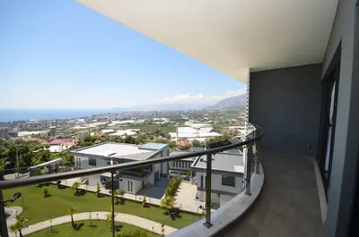 Spacious Modern Balcony with Panoramic Sea and City Views. A spacious, modern curved balcony with light gray tiled flooring and a glass-paneled railing, offering panoramic views of the sea, city, and lush green gardens below.