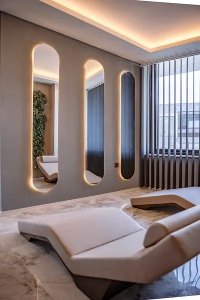 Elegant Relaxation Space with Modern Loungers and Ambient Lighting. Modern relaxation room with two elegant chaise lounges on a polished floor. Ambient backlit arched mirrors, cove lighting, and a reflected vertical garden create a serene, luxurious atmosphere.
