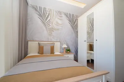 Modern Single Bedroom with Palm Leaf Wallpaper. A modern bedroom with a single bed, stylish palm leaf wallpaper, a white wardrobe with a mirrored door, and a bedside table. The room is well-lit and features sheer curtains.
