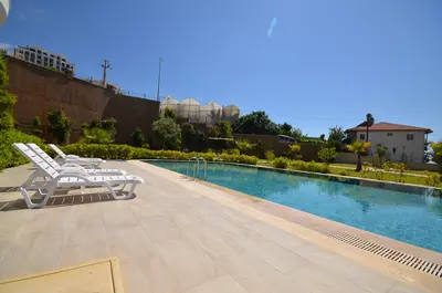 Inviting Outdoor Swimming Pool with Sun Loungers and Garden. A long, modern outdoor swimming pool with a clean paved deck and two white lounge chairs. Surrounded by lush green landscaping under a bright blue sky, ideal for relaxation.