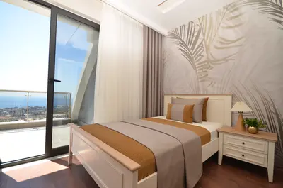 Modern Bedroom with Balcony Access and Panoramic Sea View. Modern bedroom with dark wood floor and tropical wallpaper. A large glass door opens to a balcony, offering panoramic sea and city views with natural light.