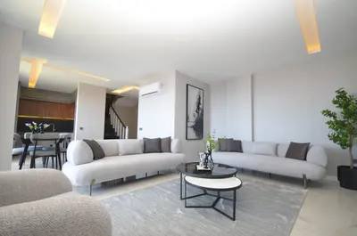 Spacious Contemporary Living Area with Stylish Furnishings. A modern, spacious living room with light grey sofas, nested coffee tables, and a soft grey rug. Contemporary ceiling lights and air conditioning are visible, opening to a dining area.