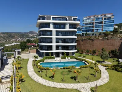 Modern Apartment Building with Private Pool and Landscaped Garden. Exterior view of a modern apartment complex featuring a multi-story building, a sparkling swimming pool, and a beautifully landscaped garden with palm trees and a paved path, set against a mountain backdrop.