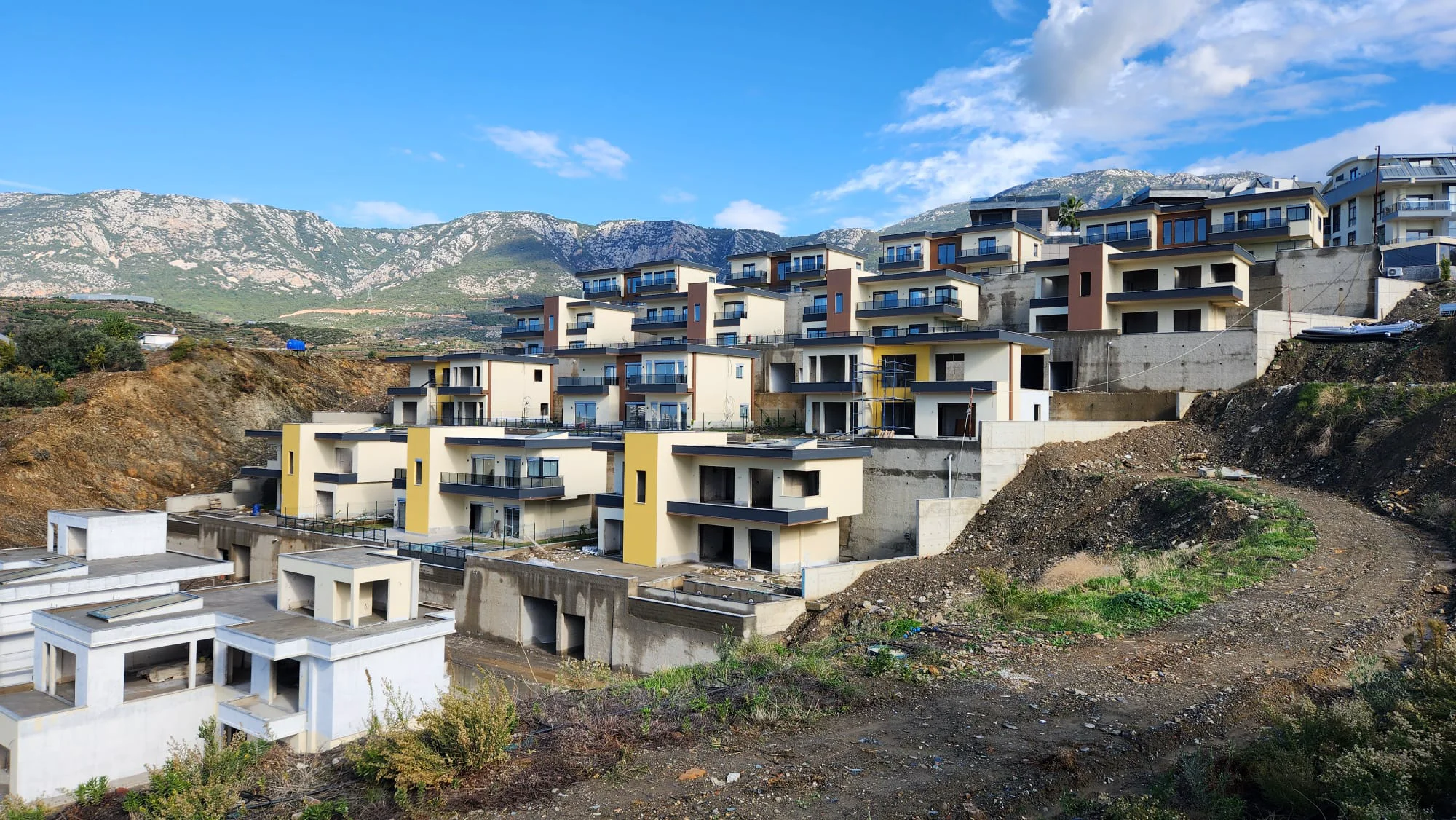 New Hillside Residential Complex with Mountain Views. Exterior view of a modern residential complex under construction on a steep hillside. Multiple terraced villas with balconies offer mountain views under a blue sky.