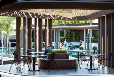 Elegant Outdoor Social Area with Poolside Lounge. An elegant outdoor social area featuring modern black chairs and marble-top tables under a decorative pergola. A sparkling swimming pool, lush palm trees, and people relaxing create a luxurious resort-like atmosphere.