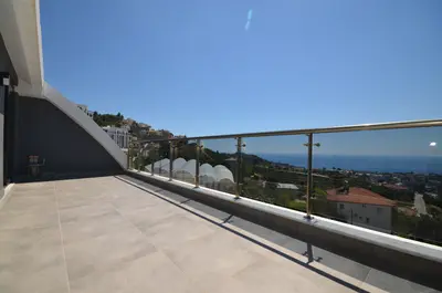 Modern Balcony with Panoramic Sea and City Views. This modern balcony features light tile flooring and a glass railing, offering stunning panoramic views of a hillside town and the sparkling blue Mediterranean Sea.