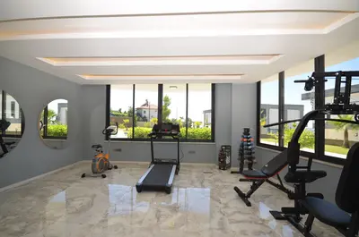 Modern Home Gym with Pool and Garden Views. A modern home gym featuring a treadmill, exercise bike, and weights. It has polished marble flooring, large windows, recessed lighting, and offers a view of the pool and garden.