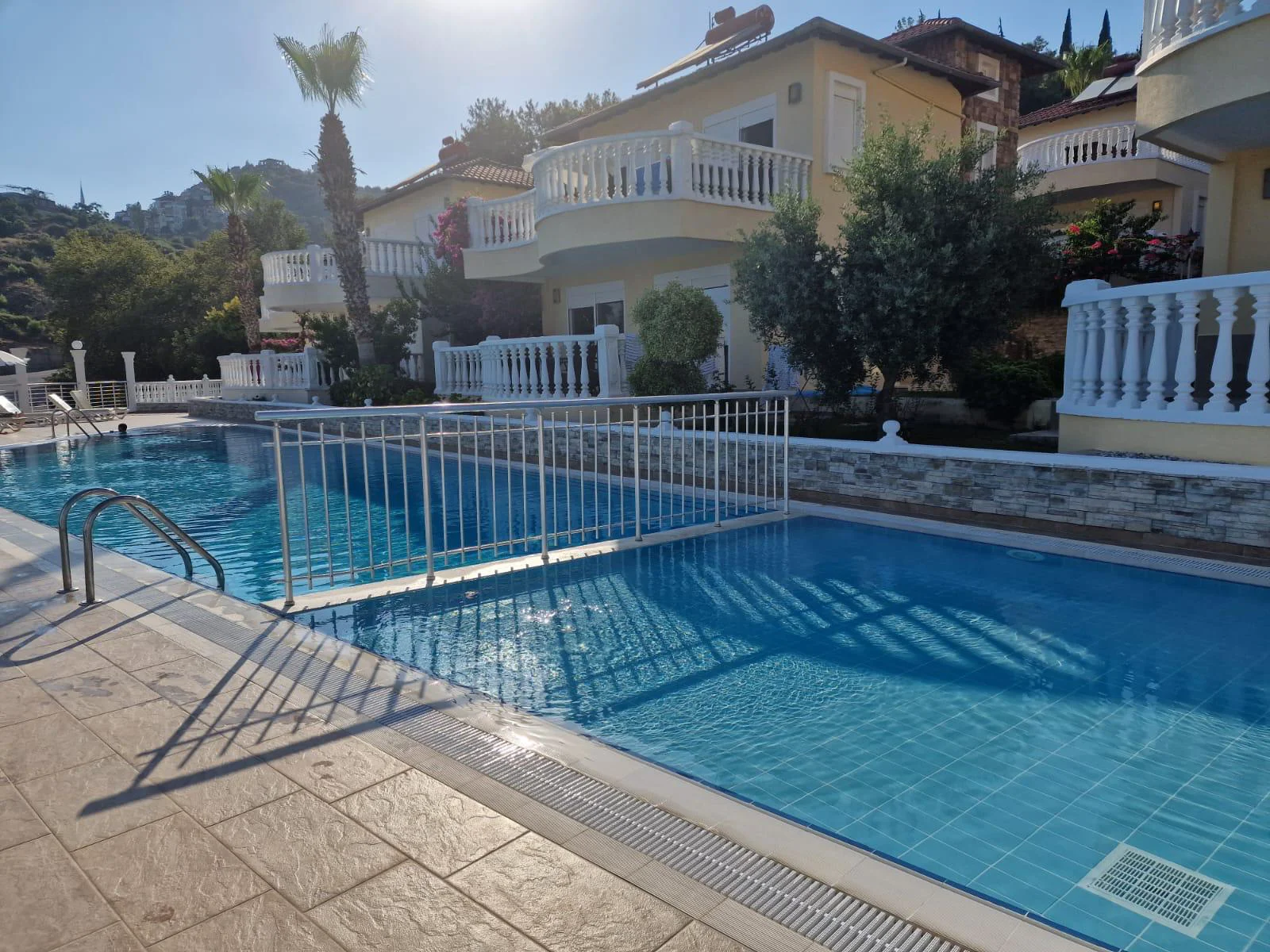 Beeindruckender Außenpoolbereich in einer mediterranen Villenanlage. Ein Außenpoolbereich mit zwei hellblauen, gefliesten Pools, die durch einen weißen Zaun getrennt sind. Das moderne Anwesen umfasst eine geflieste Terrasse, üppiges Grün und Villen in sonniger mediterraner Umgebung.