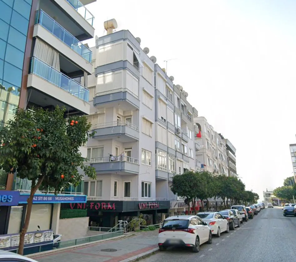 Modernes Mehrfamilienhaus von außen mit Stadtstraßenansicht. Außenansicht eines modernen Mehrfamilienhauses an einer Stadtstraße. Es verfügt über mehrere Balkone, eine weiß-graue Fassade und Gewerbeflächen im Erdgeschoss. Geparkte Autos säumen die von Bäumen gesäumte Straße.