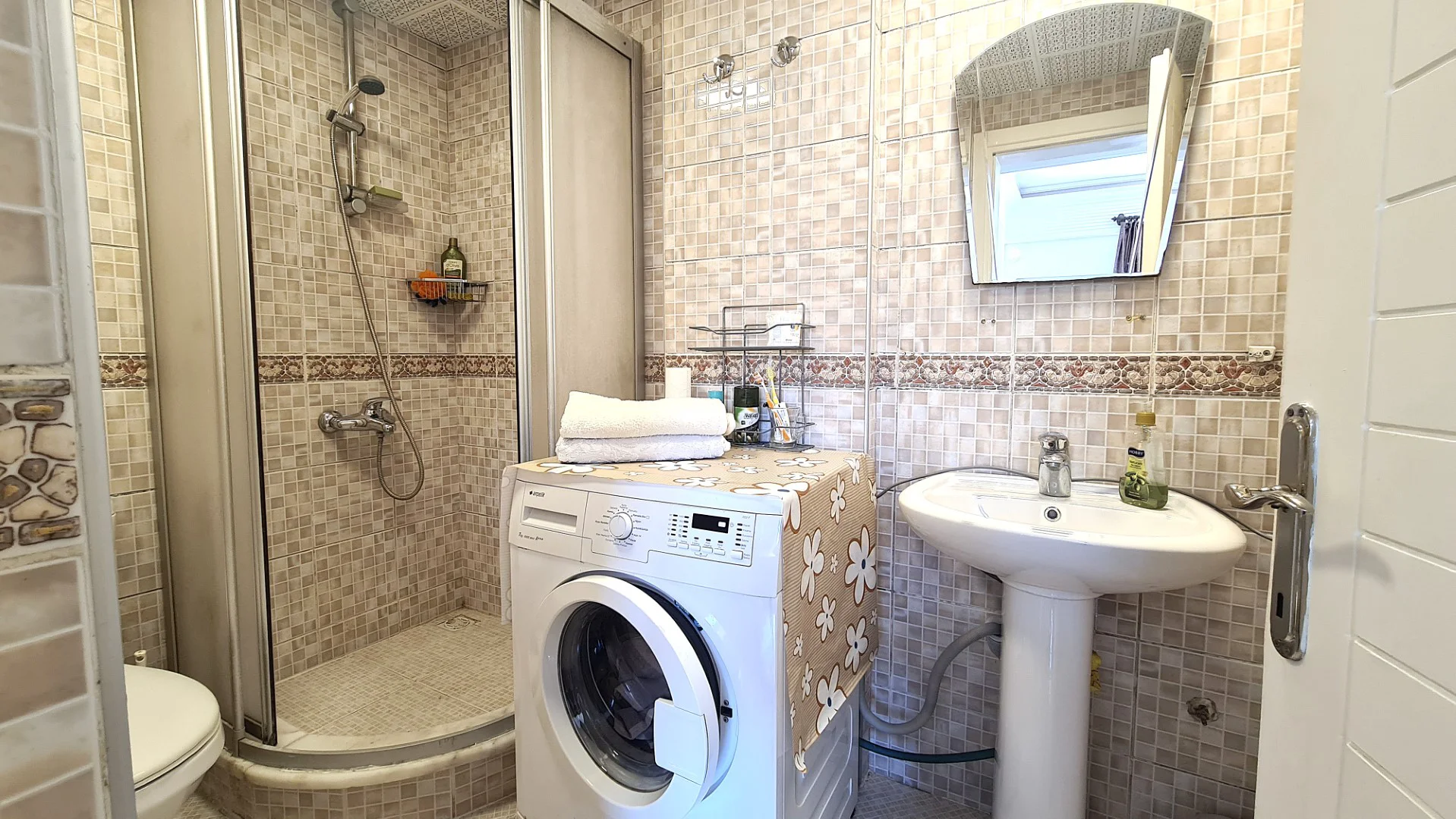 Funktionales Badezimmer mit Duschkabine und Waschmaschine. Ein gepflegtes Badezimmer mit Wänden in Mosaikoptik, einer geräumigen Duschkabine und einem modernen Säulenwaschbecken. Enthält eine praktische Waschmaschine für zusätzlichen Komfort.
