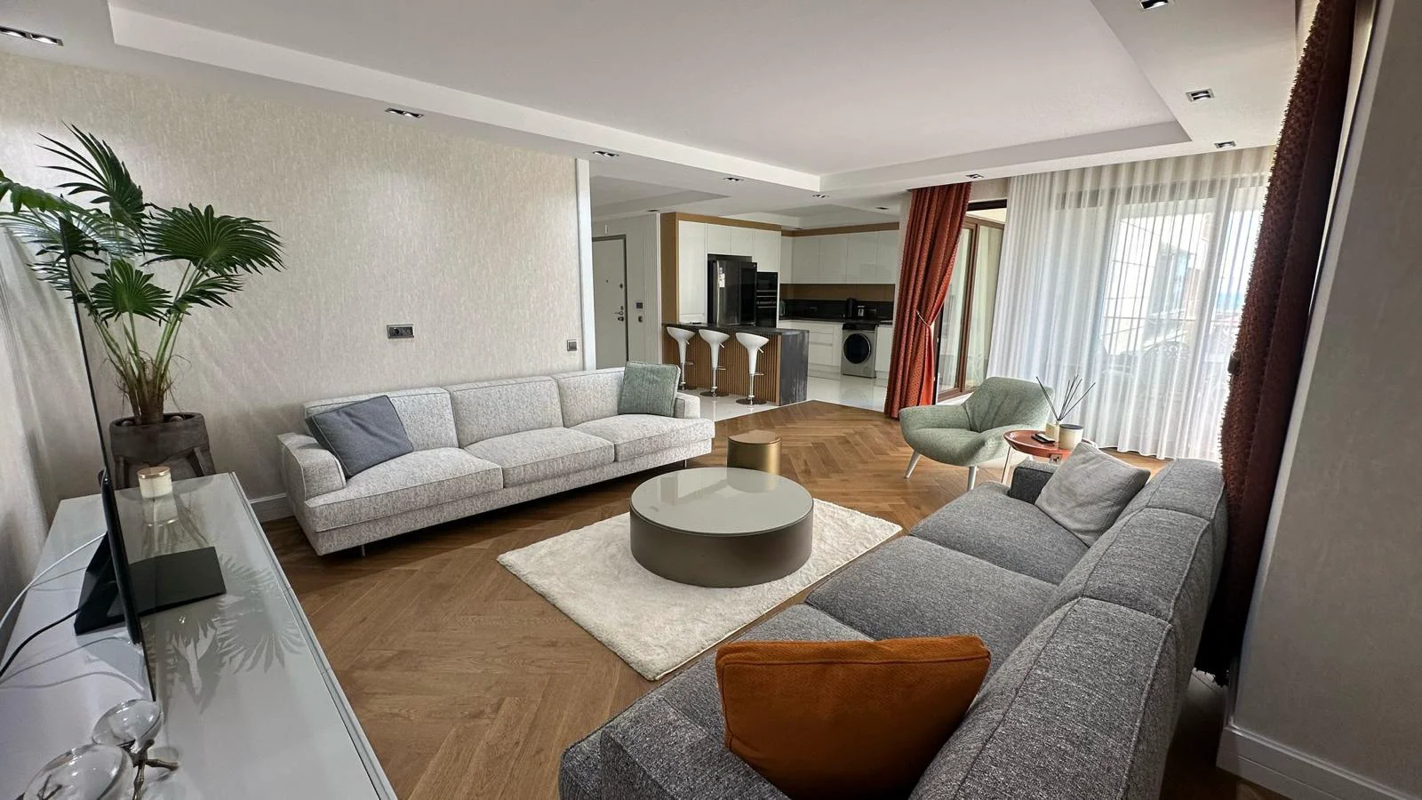 Geräumiges offenes Wohnzimmer mit moderner Küche. Ein modernes offenes Wohnzimmer mit Fischgrät-Parkettböden, zwei bequemen Sofas und einem runden Couchtisch. Der moderne Küchenbereich verfügt über eine Kochinsel, moderne Geräte und große Fenster.