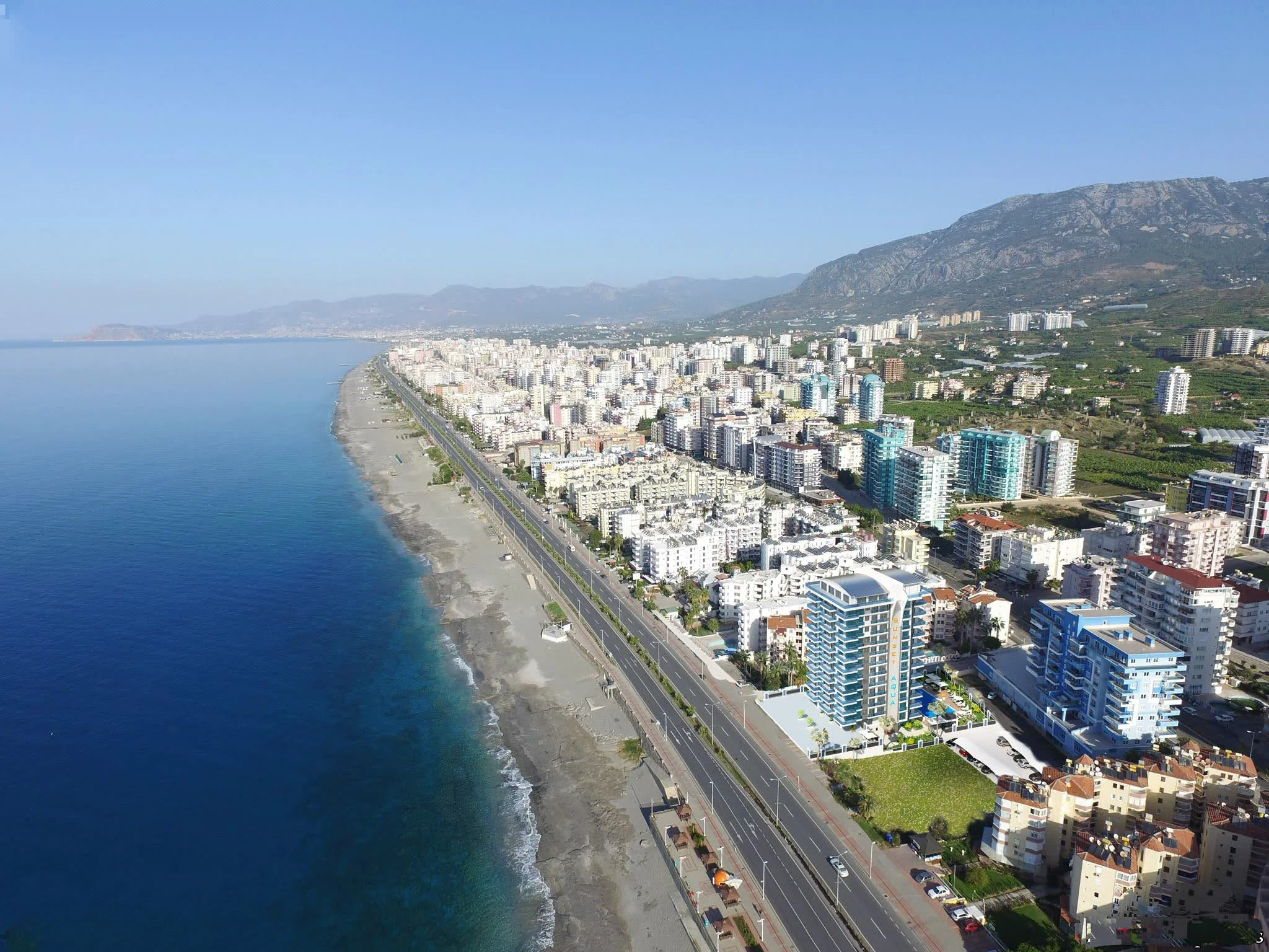 Luftaufnahme der Küstenstadt Alanya mit Strand und Bergen. Luftaufnahme einer lebhaften Küstenstadt in der Türkei mit modernen Gebäuden entlang eines langen Sandstrandes und dem blauen Mittelmeer, mit majestätischen Bergen im Hintergrund.