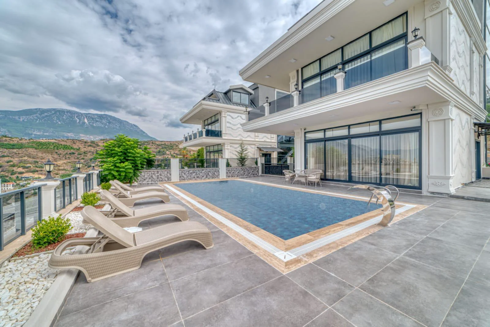 Moderne Villa – Außenpoolbereich mit Bergblick. Ein moderner Außenpoolbereich mit einem klaren blauen Swimmingpool, bequemen Liegestühlen und einer Essgruppe im Freien. Das renovierte Anwesen besticht durch einen atemberaubenden Blick auf die Berglandschaft unter einem bewölkten Himmel.