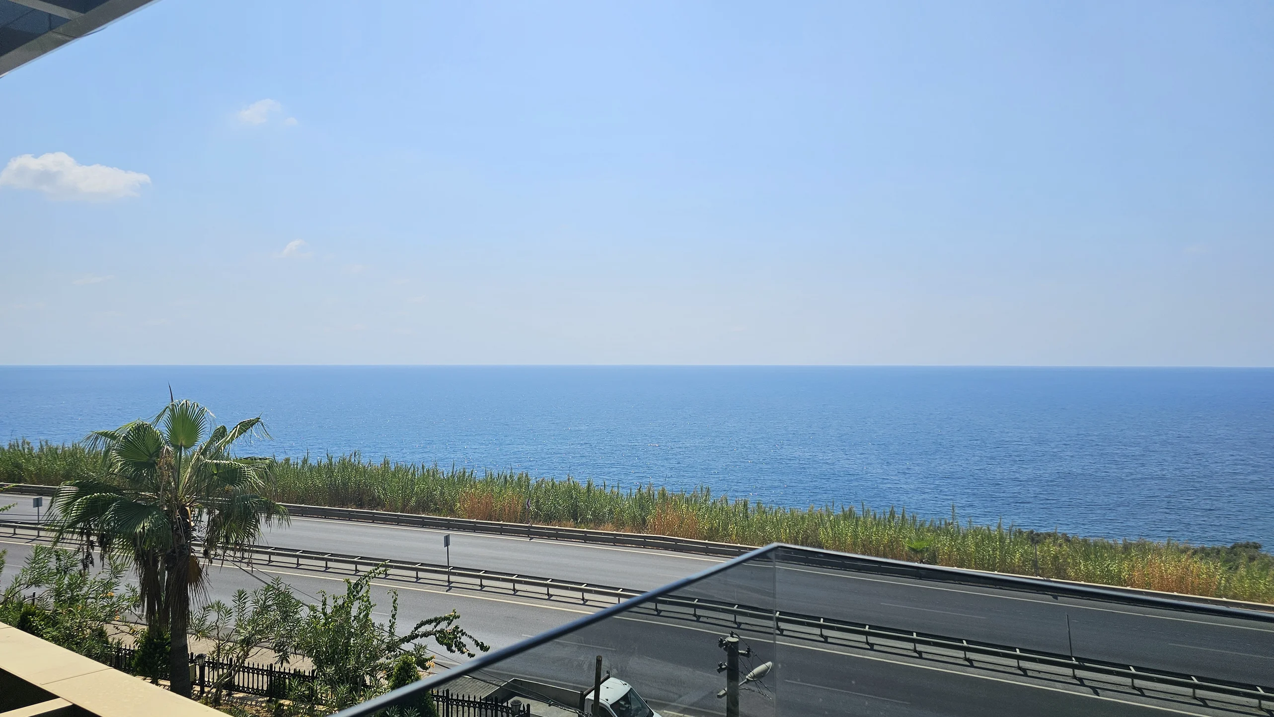 Atemberaubender Meerblick vom Balkon mit Küstenstraße. Ein heller, malerischer Blick von einem modernen Balkon auf das tiefblaue Mittelmeer und eine Küstenautobahn, mit einer üppig grünen Palme im Vordergrund.