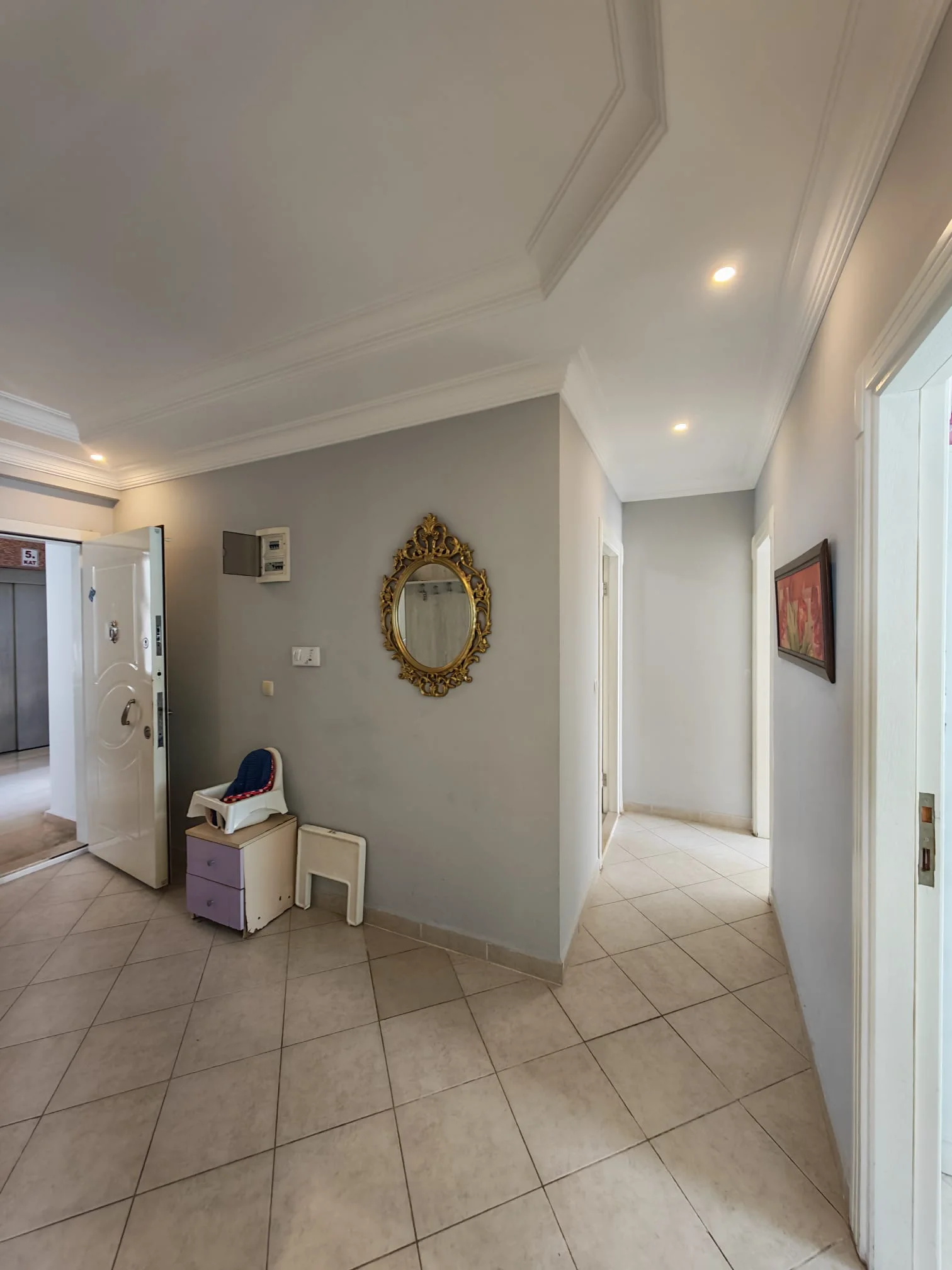Moderner Eingangsbereich einer Wohnung mit Fliesenboden. Der helle Flur einer Wohnung besticht durch hellgraue Wände, moderne Einbaustrahler und einen beigen Fliesenboden. Ein verzierter goldener Spiegel an der Wand verleiht dem modernen Raum eine klassische Note.