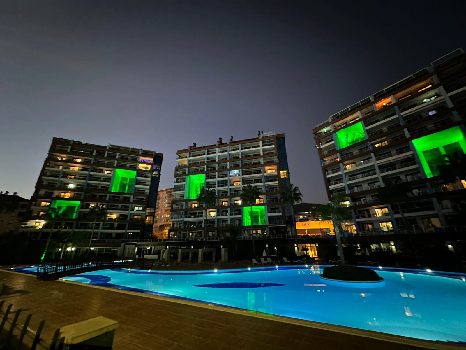 Moderner Wohnkomplex mit beleuchtetem Pool bei Nacht. Außenansicht eines modernen Wohnkomplexes bei Nacht mit einem großen, hell beleuchteten Swimmingpool und drei zeitgenössischen Wohngebäuden mit sichtbaren Balkonen und grün beleuchteten Fenstern.