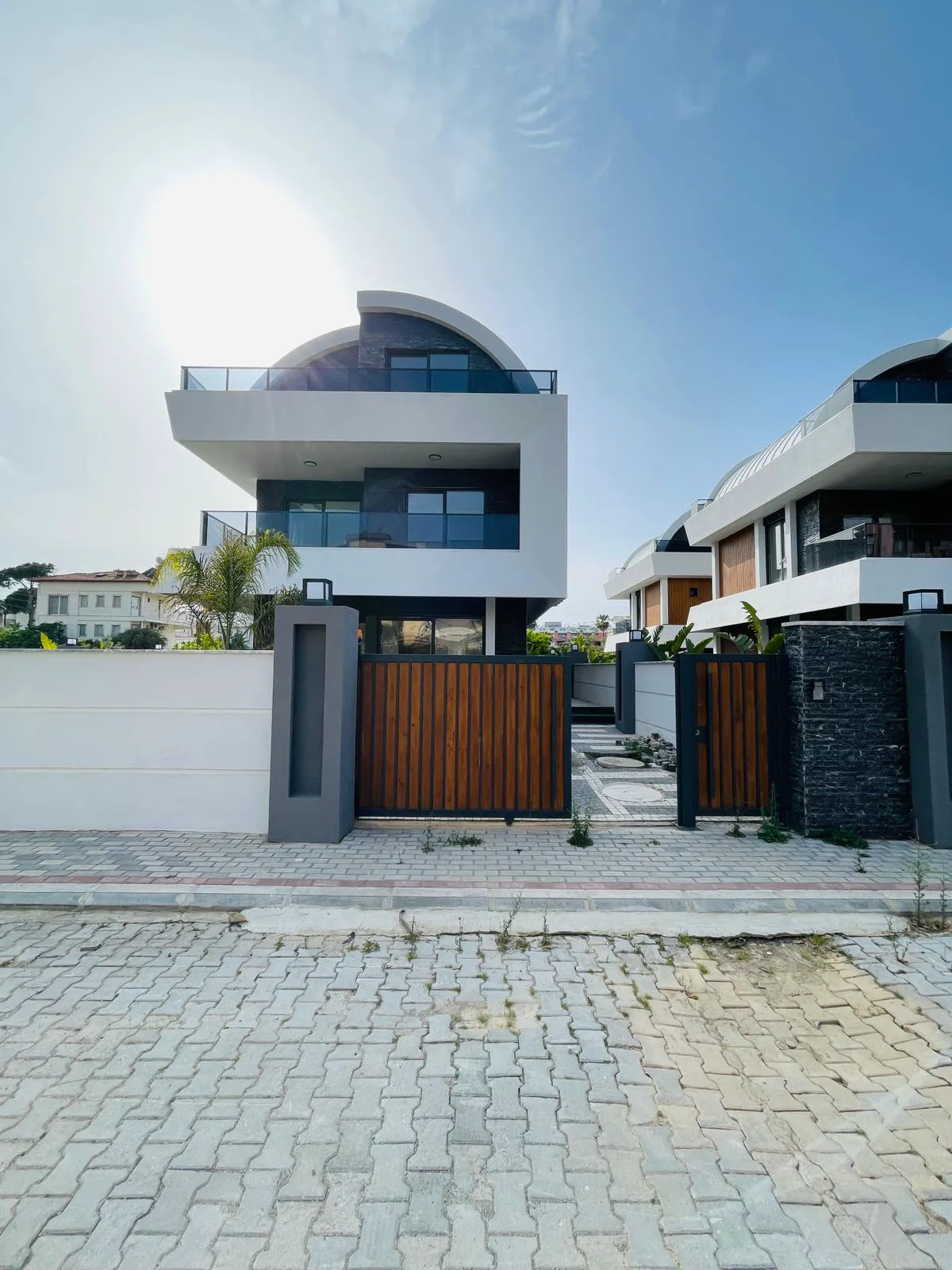 Moderne Villa-Außenansicht mit zeitgenössischem Design und Eingangsbereich. Moderne Außenansicht einer Villa mit markantem geschwungenem Dach, weiß-dunkelgrauer Fassade und Glasbalkonen. Ein stilvolles Holztor führt zum gepflasterten Privateingang und Garten.