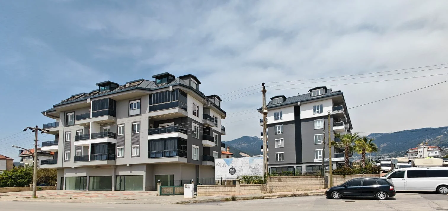 Moderne Apartmentgebäude Außenansicht mit Bergblick. Außenansicht von zwei modernen, neu gebauten Apartmentgebäuden mit grau-weißen Fassaden, Balkonen und großen Fenstern. Straßenszene mit geparkten Autos und fernen Bergen unter bewölktem Himmel.
