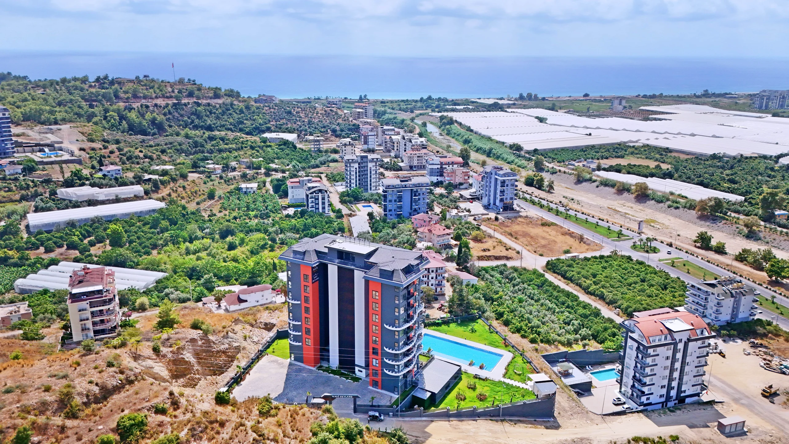 Küstennaher Apartmentkomplex mit Pool und Meerblick in der Türkei. Luftaufnahme einer modernen Wohnanlage in einer üppigen Küstenregion der Türkei. Zu den Merkmalen gehören zeitgenössische Gebäude, ein klarer Swimmingpool, grüne Außenanlagen sowie Fernblick auf das Meer und die umliegenden landwirtschaftlichen Flächen.