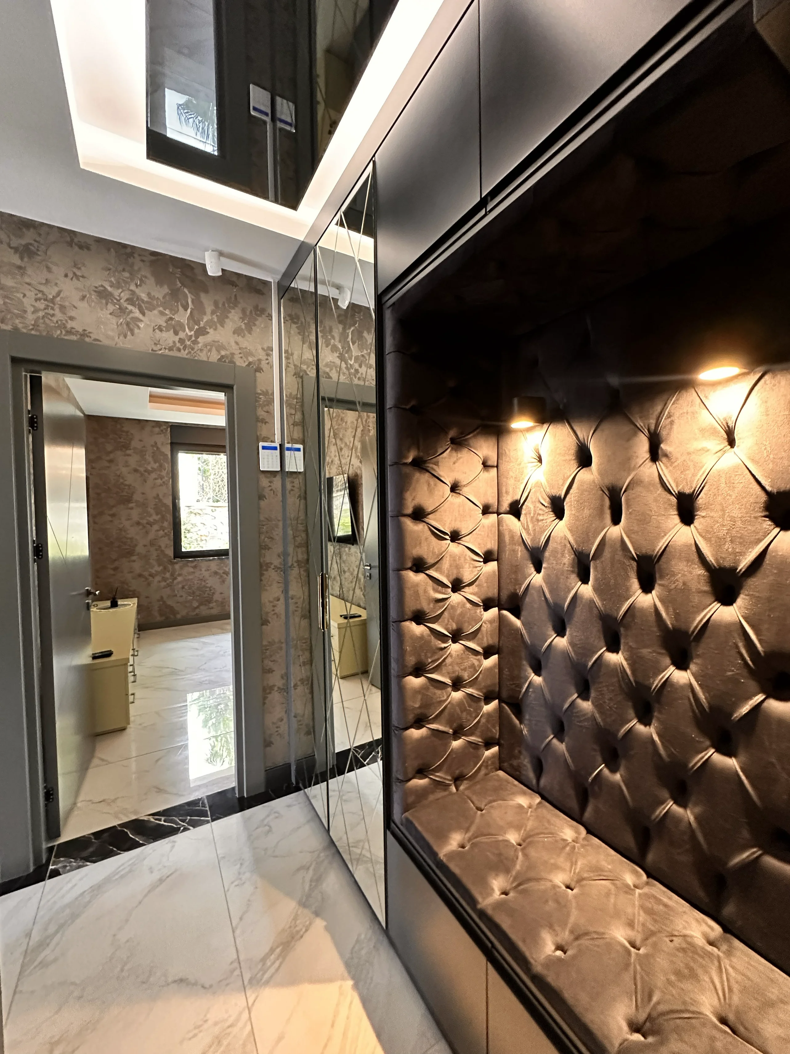 Luxuriöser Eingangsbereich mit maßgefertigter getufteter Sitzgelegenheit und verspiegeltem Kleiderschrank. Ein moderner, luxuriöser Eingangsbereich mit einer eingebauten getufteten Samtsitzbank und Strahlern. Verspiegelte Kleiderschranktüren und weißer Marmoroptik-Boden schaffen einen eleganten und funktionalen Raum.