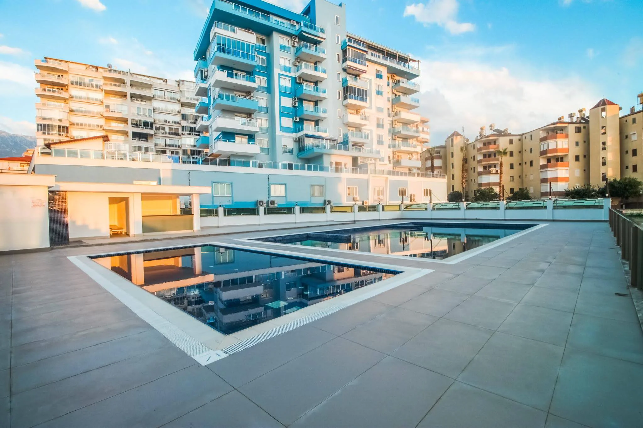 Wohnanlage mit glitzernden Außenpools. Außenbereich einer modernen Wohnanlage in der Türkei mit zwei makellosen rechteckigen Swimmingpools auf einer gefliesten Terrasse. Umgebende Wohngebäude und klarer blauer Himmel.