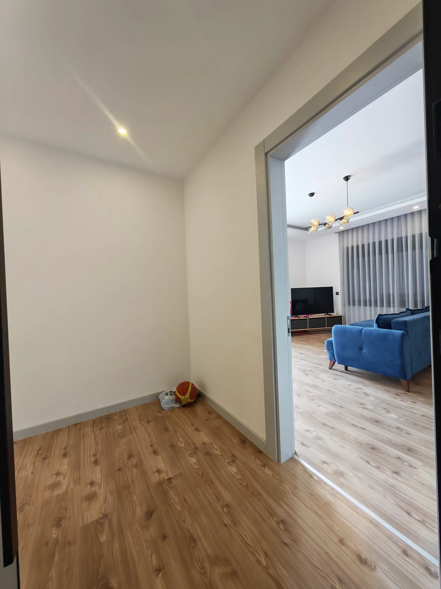 Moderner Flurübergang zum großzügigen Wohnzimmer. Moderner Flur mit Laminatboden in Holzoptik, der in ein helles Wohnzimmer führt. Zur Ausstattung gehören ein blaues Sofa, ein großer Fernseher, ein stilvoller Kronleuchter und weiße Wände.