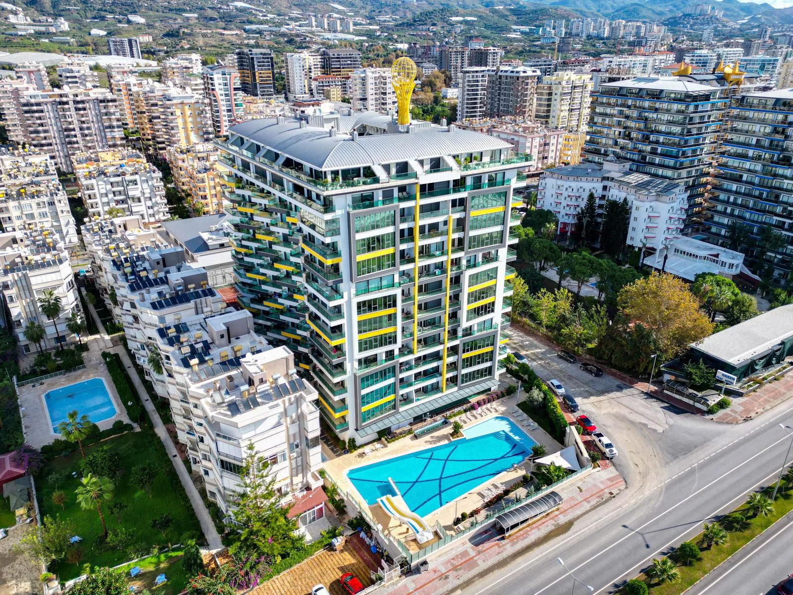 Moderner Apartmentkomplex mit Außenpool und Stadtblick. Luftaufnahme eines modernen Apartmentkomplexes mit einem großen Außenpool und einer Rutsche. Das Gebäude mit gelben und grauen Akzenten liegt in einem lebendigen Stadtbild mit Bergen in der Ferne.