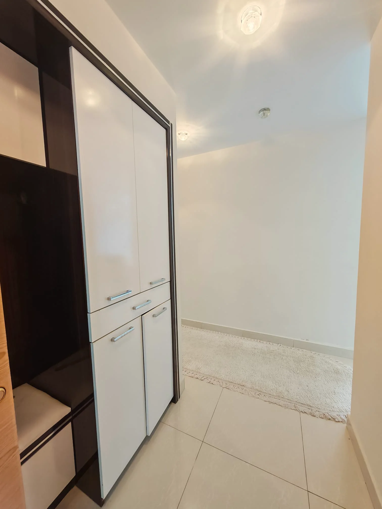 Moderner Flur-Eingangsbereich mit Einbauschrank und Sitzgelegenheit. Ein sauberer, moderner Eingangsbereich mit einem weißen Einbauschrank mit silbernen Griffen und einer kleinen Polsterbank. Der Raum verfügt über helle Fliesenböden und Deckeneinbauleuchten.