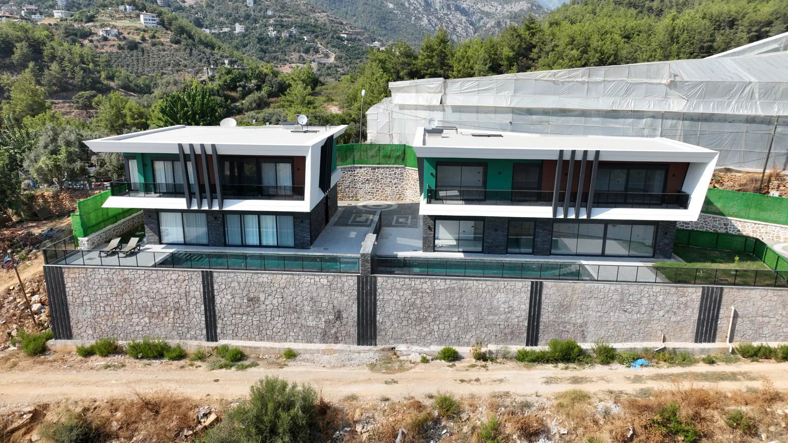 Luxuriöse moderne Villen mit privaten Pools und Bergblick. Luftaufnahme von zwei modernen Luxusvillen mit privaten Infinity-Pools, großen Fenstern und Balkonen. Auf einem üppigen grünen Hügel gelegen, bieten diese zeitgenössischen Häuser einen atemberaubenden Blick auf die Berge.