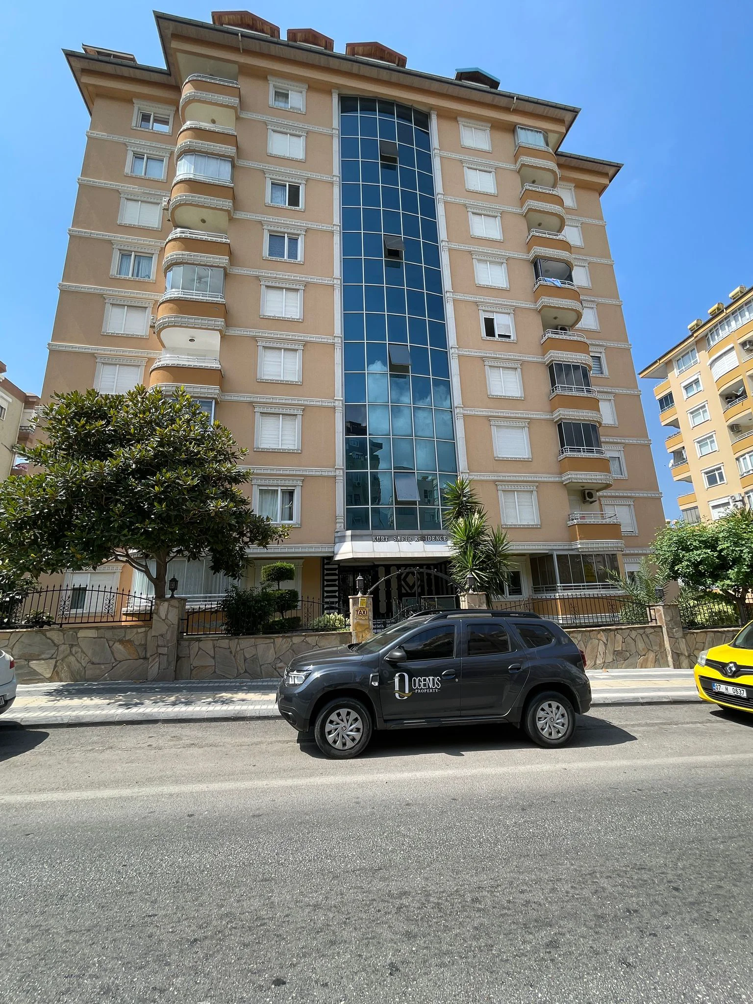 Moderne Wohnhaus-Fassade mit Balkonen. Außenansicht eines modernen mehrstöckigen Wohnhauses mit beiger Fassade, weißen Fenstern, geschwungenen Balkonen und einem zentralen blauen Glaselement an einer sonnigen Straße.