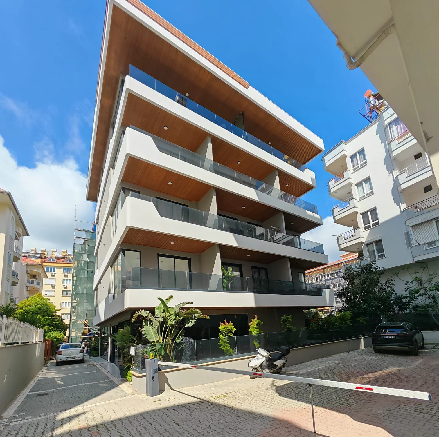 Moderne Außenansicht eines Mehrfamilienhauses mit Glasbalkonen. Moderne Außenansicht eines Mehrfamilienhauses mit zeitgenössischer Architektur, Glasbalkonen und Decken in Holzoptik. Das Anwesen verfügt über eine gesicherte Parkhauseinfahrt und grüne Außenanlagen.