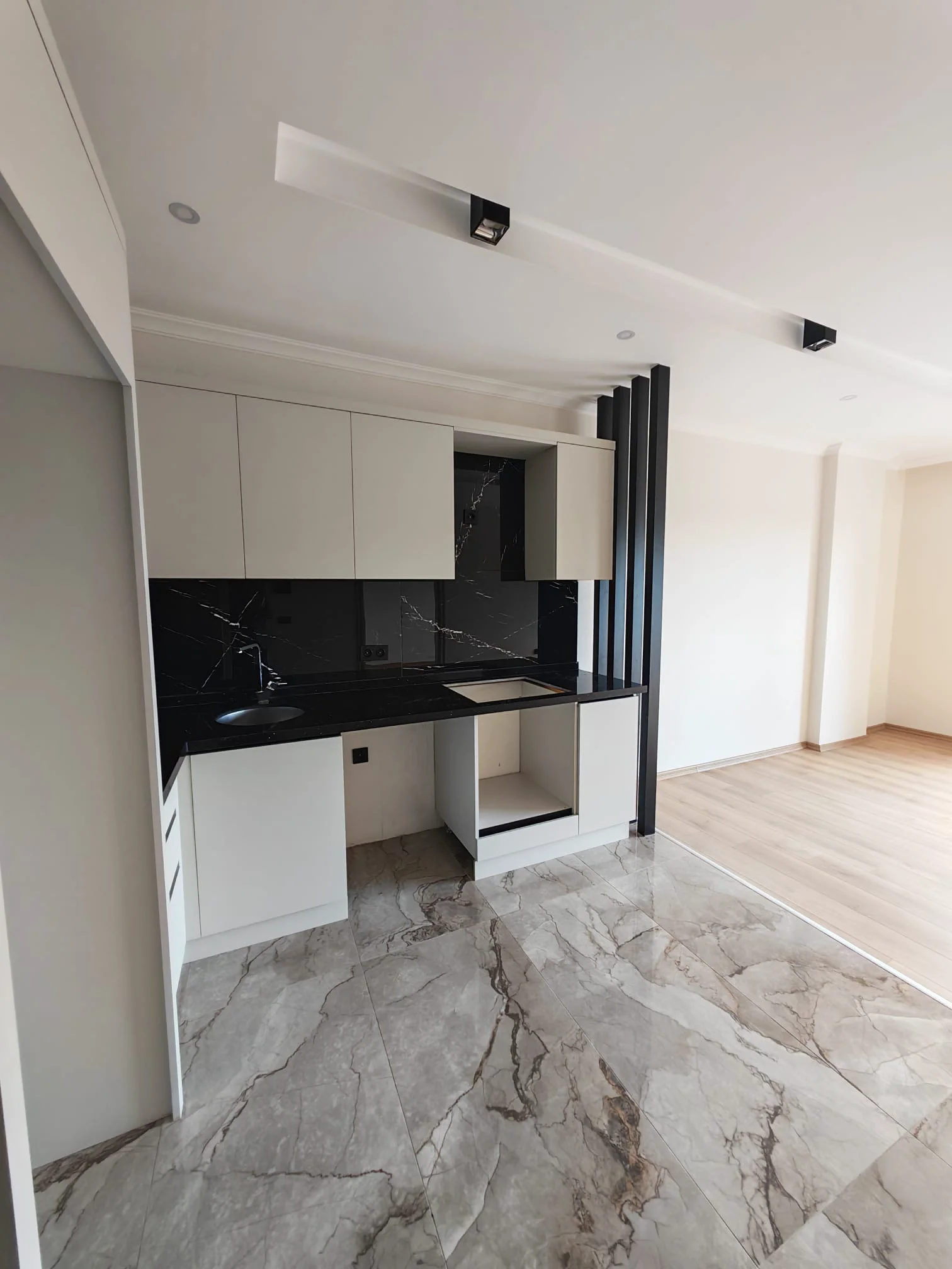 Moderne offene Küche mit Marmorakzenten. Eine moderne, frisch renovierte offene Küche mit weißen Schränken, schwarzem Marmorspiegel und grauem Fliesenboden. Schienenbeleuchtung und ein dekorativer Raumteiler unterstreichen das zeitgemäße Design.