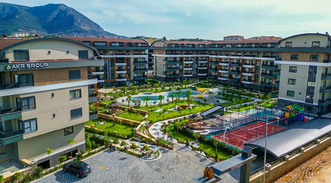 Moderner Wohnkomplex mit Pool und Bergblick. Luftaufnahme eines modernen Wohnkomplexes mit mehreren Gebäuden, einem großen Swimmingpool mit Rutschen, angelegten Gärten, einem Sportplatz und einem Kinderspielplatz vor einer Bergkulisse.